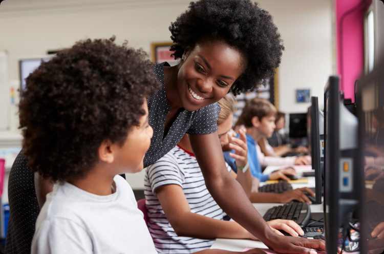 A teacher leaning in to help a student working at a computer, in a classroom of students at screens